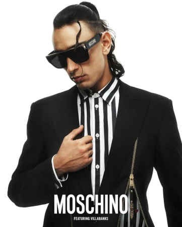Adult male poses indoors wearing a striped shirt, black jacket, and sunglasses, with a Moschino logo in white against a white background.