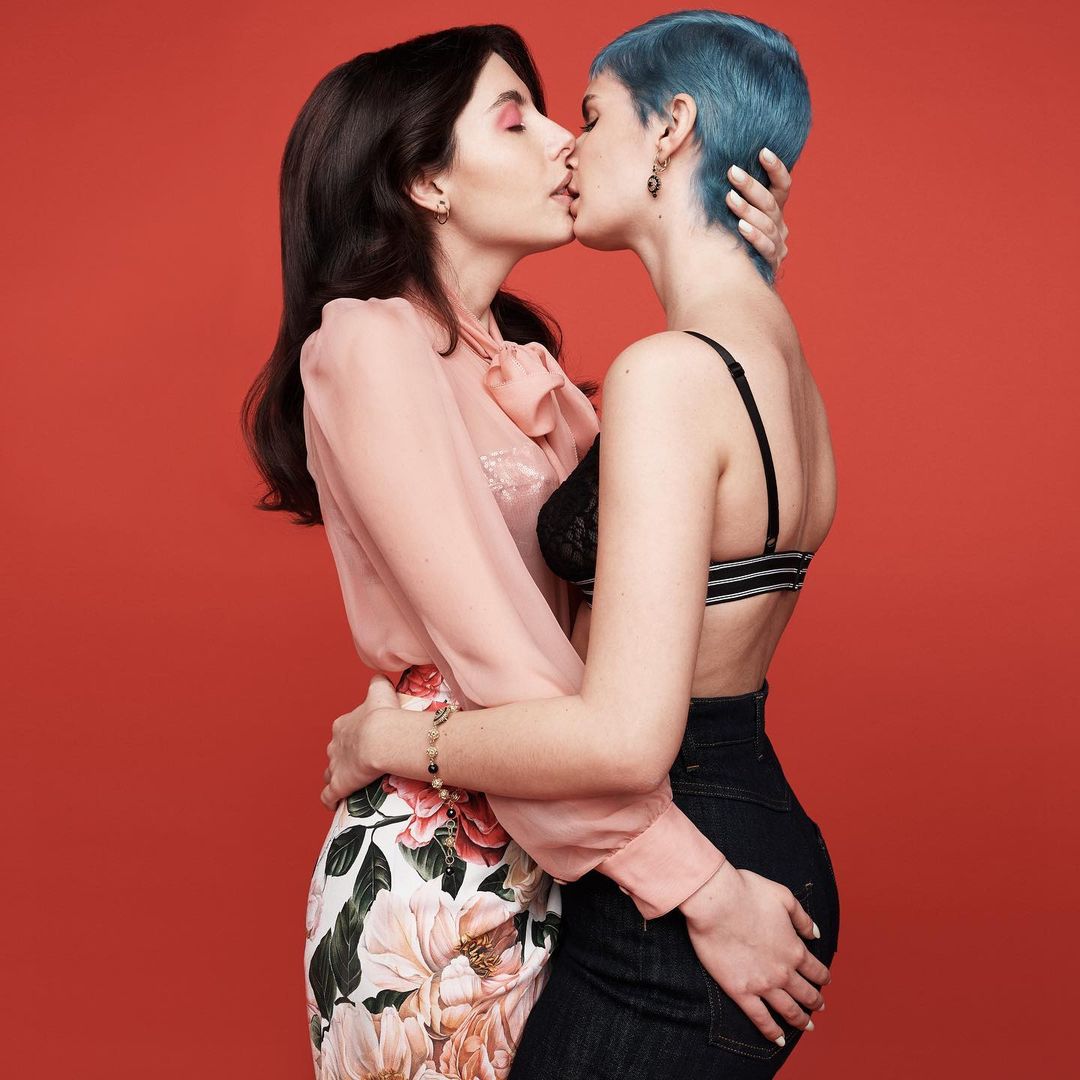 Two women kissing in front of a red background. The woman on the left has long dark hair and the woman on the right has blue hair.