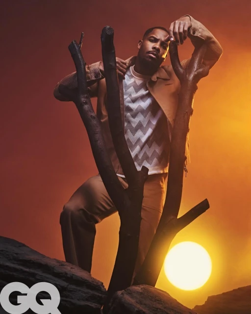 Michael B. Jordan poses in tan clothing next to a tree trunk with the sun behind him in a studio shot for GQ magazine.