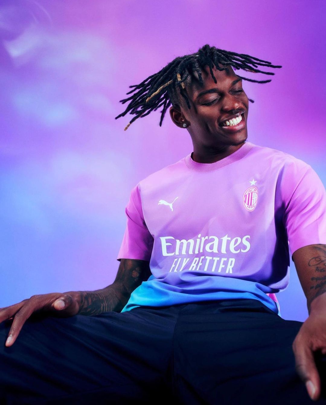 Close-up of a smiling adult male in a colorful soccer jersey against a light purple and blue background in a studio setting.