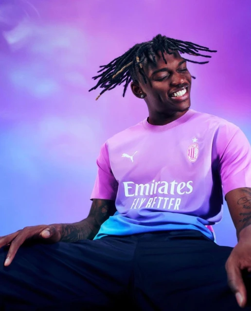 Close-up of a smiling adult male in a colorful soccer jersey against a light purple and blue background in a studio setting.