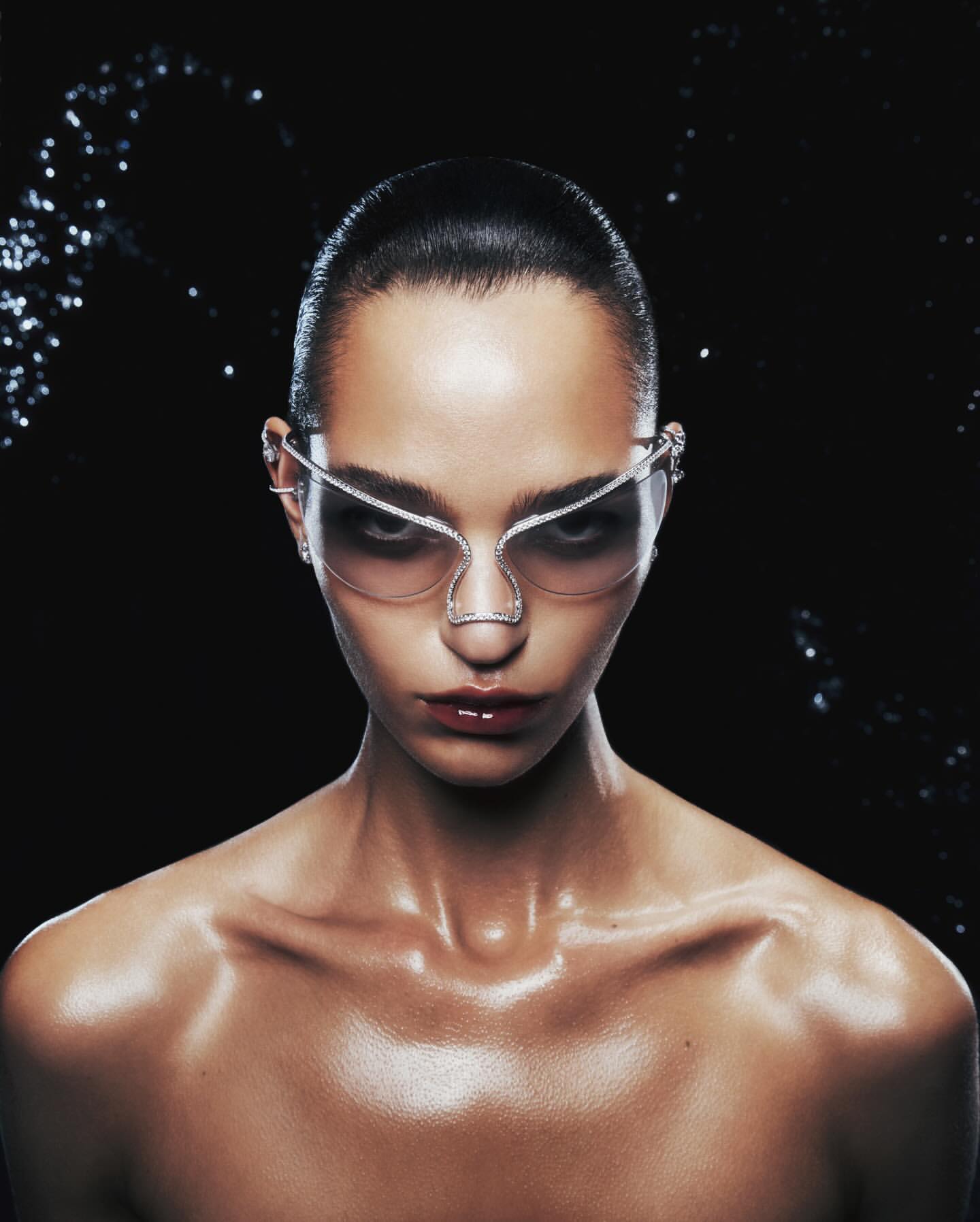 Close-up portrait of a woman wearing futuristic sunglasses with a jeweled frame against a dark background with bokeh-like highlights.