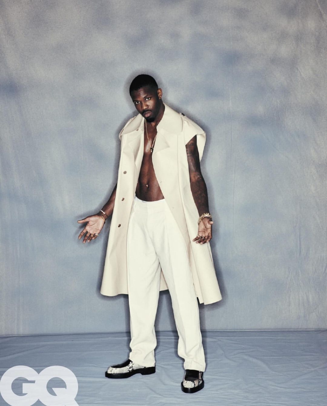An adult male is posing in a cream-colored sleeveless coat, matching pants, and gold jewelry against a grey studio backdrop with GQ branding.
