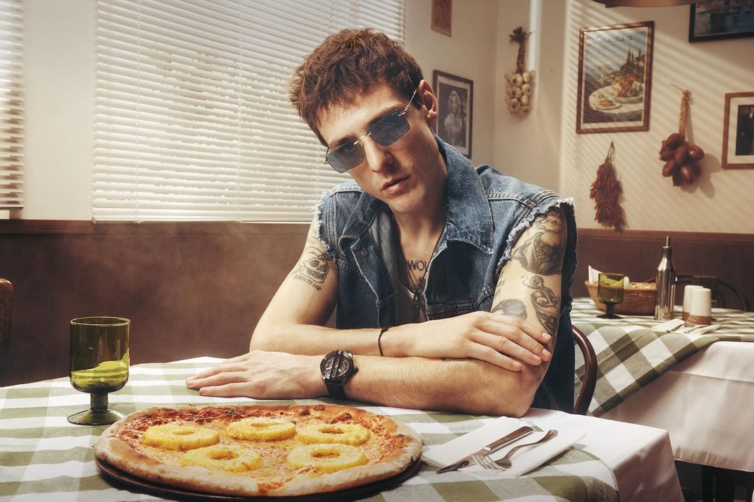 Young man with sunglasses poses at a table with pizza in an indoor setting with natural light and a checkered tablecloth.