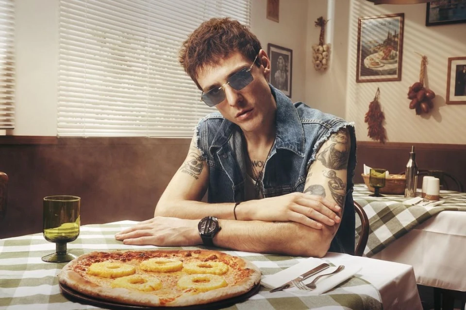 Young man with sunglasses poses at a table with pizza in an indoor setting with natural light and a checkered tablecloth.