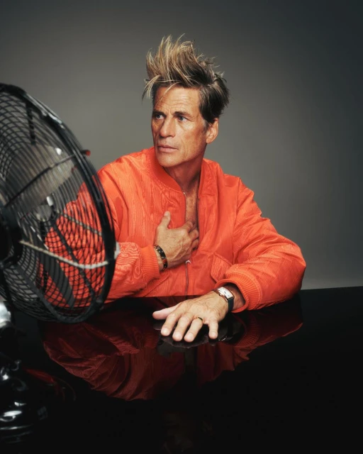 Portrait of a mature man with spiked hair, an open orange jacket, black table, and an oscillating fan in a studio setting.