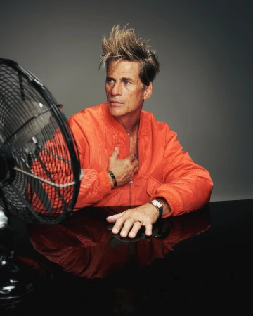 Portrait of a mature man with spiked hair, an open orange jacket, black table, and an oscillating fan in a studio setting.