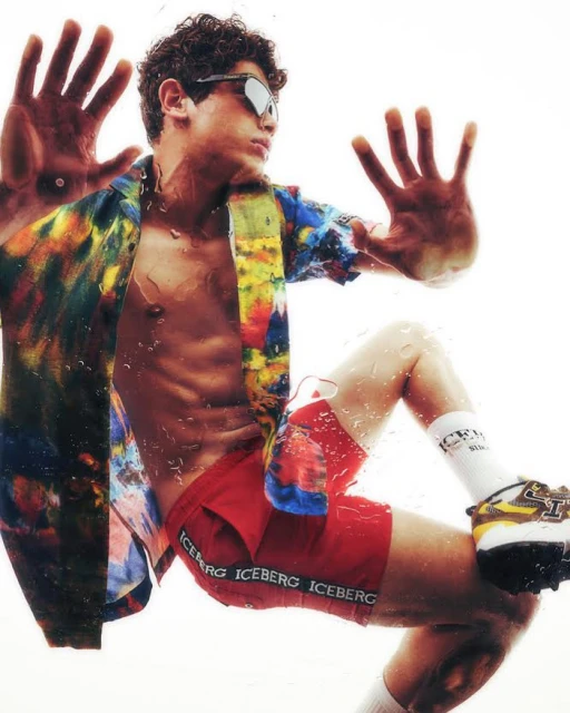 A young male model in a partially unbuttoned tie-dye shirt, red shorts, white socks, and sneakers poses with his hands up against glass.