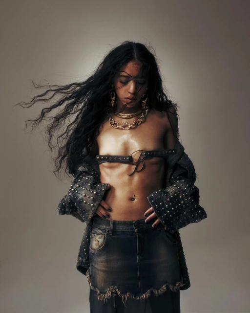 Adult female with long black hair wearing a denim skirt, studded jacket, and gold chains posing in a photo studio with a neutral background.