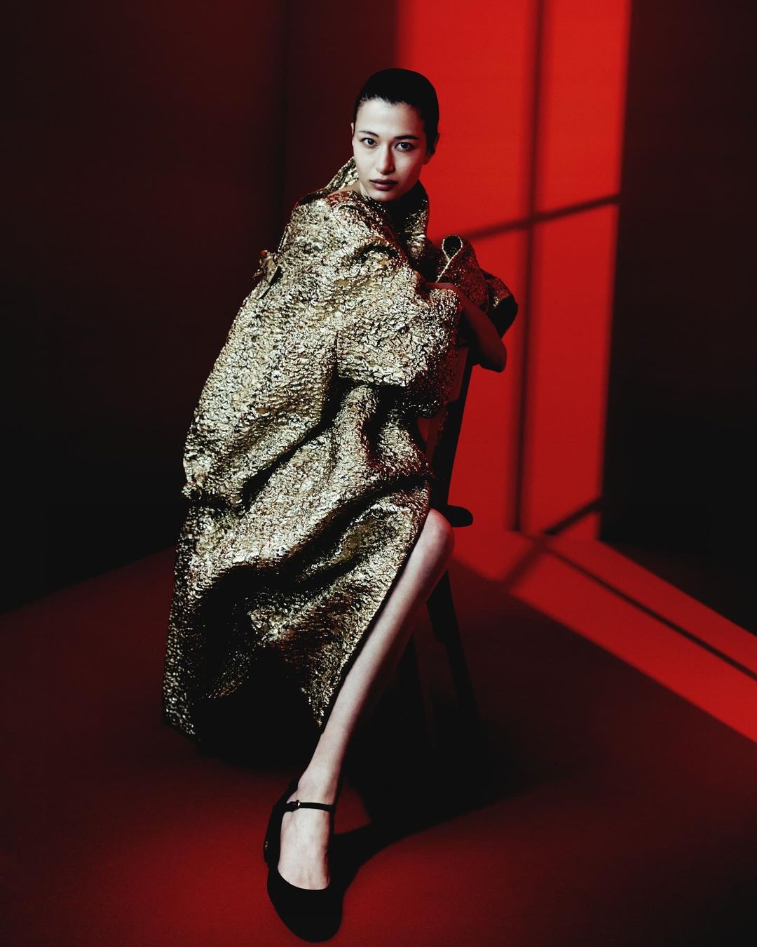 Woman poses in a shimmering gold coat in a red room, seated on a chair, showcasing a fashion-forward look in a studio setting.