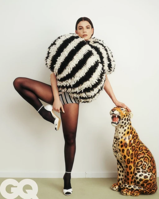Woman poses in striped top and black tights, one leg raised, next to a ceramic jaguar against a white backdrop in a fashion editorial.