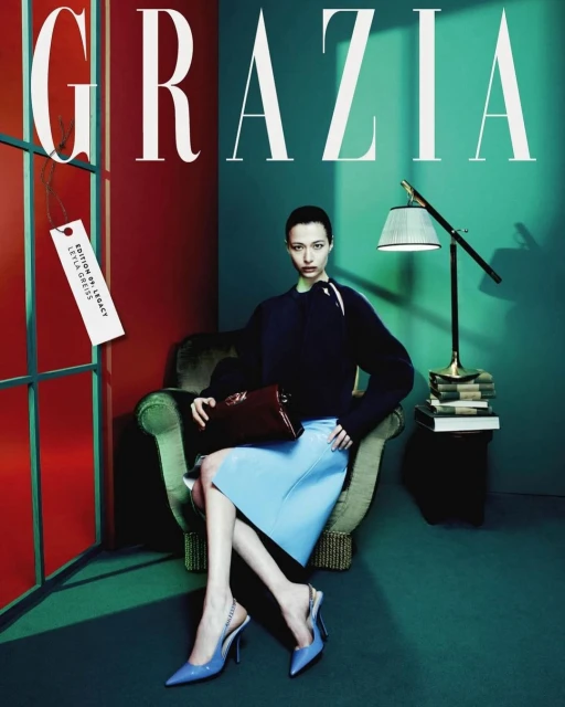 Fashion magazine cover featuring a stylish woman in a teal and red setting, seated in a green chair holding a maroon handbag.
