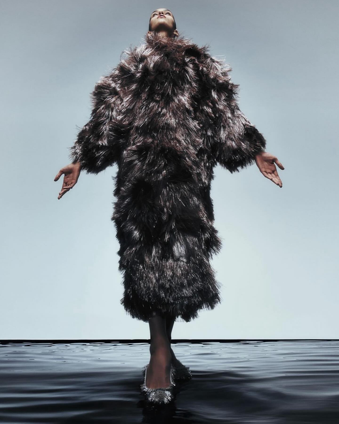 Fashionable woman in full-length feathered coat poses with arms outstretched on a glossy black surface against a gradient gray backdrop.