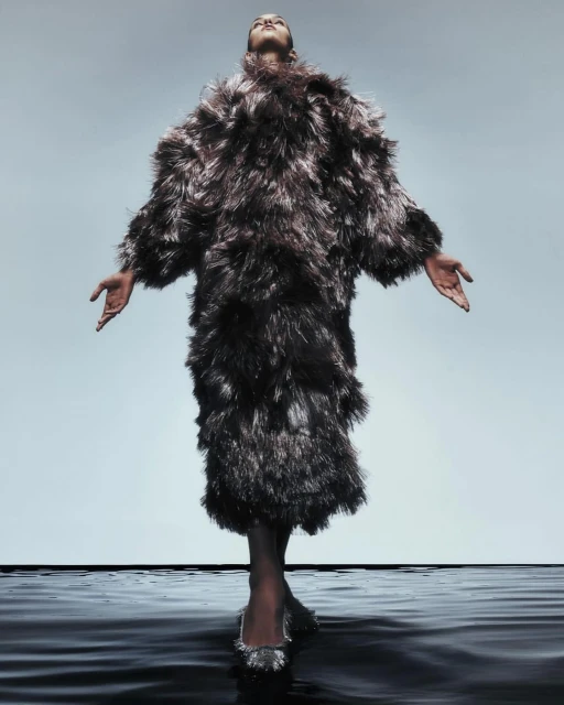 Fashionable woman in full-length feathered coat poses with arms outstretched on a glossy black surface against a gradient gray backdrop.