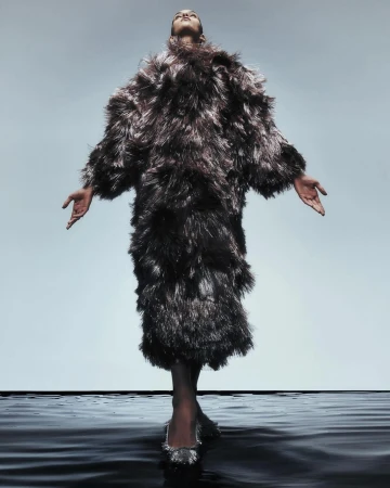 Fashionable woman in full-length feathered coat poses with arms outstretched on a glossy black surface against a gradient gray backdrop.