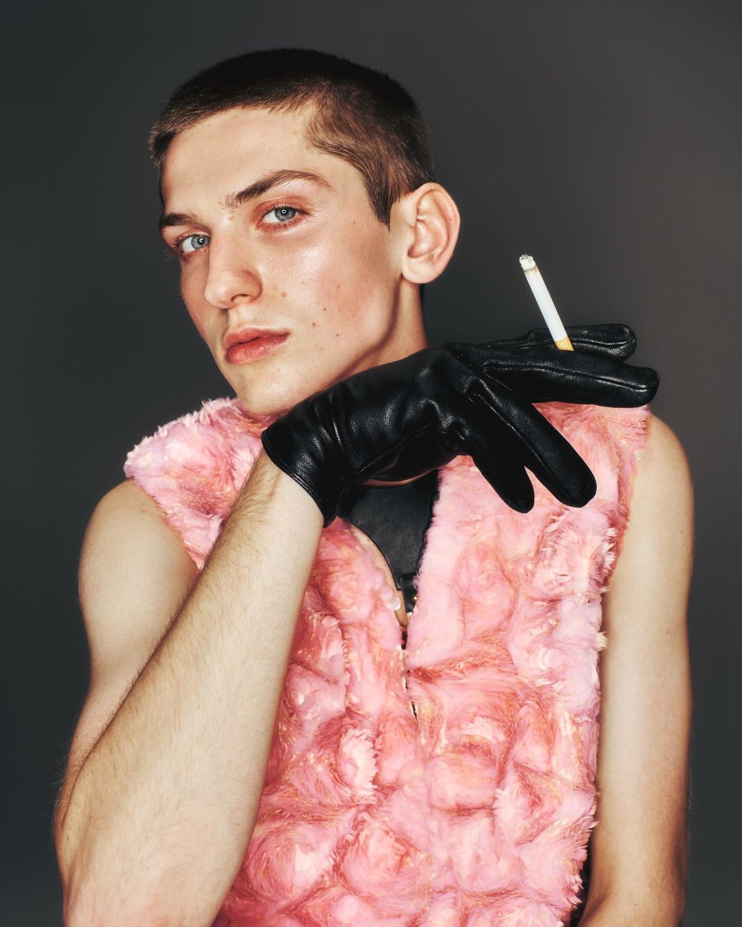 Portrait of a young adult man with black gloves and a pink faux fur vest holding a cigarette in a studio setting with a grey background.