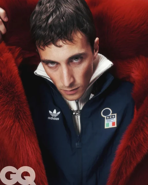 Close-up studio portrait of a young adult male wearing an Adidas tracksuit top and a red fur coat, captured at eye-level with GQ logo.