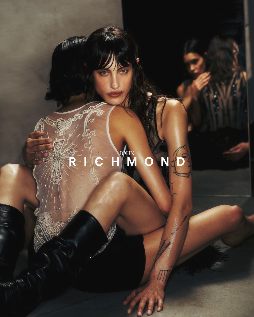 Two women embracing intimately in a dark studio setting wearing black boots and sheer embroidered tops with tattoos visible.