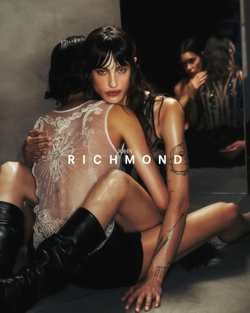 Two women embracing intimately in a dark studio setting wearing black boots and sheer embroidered tops with tattoos visible.