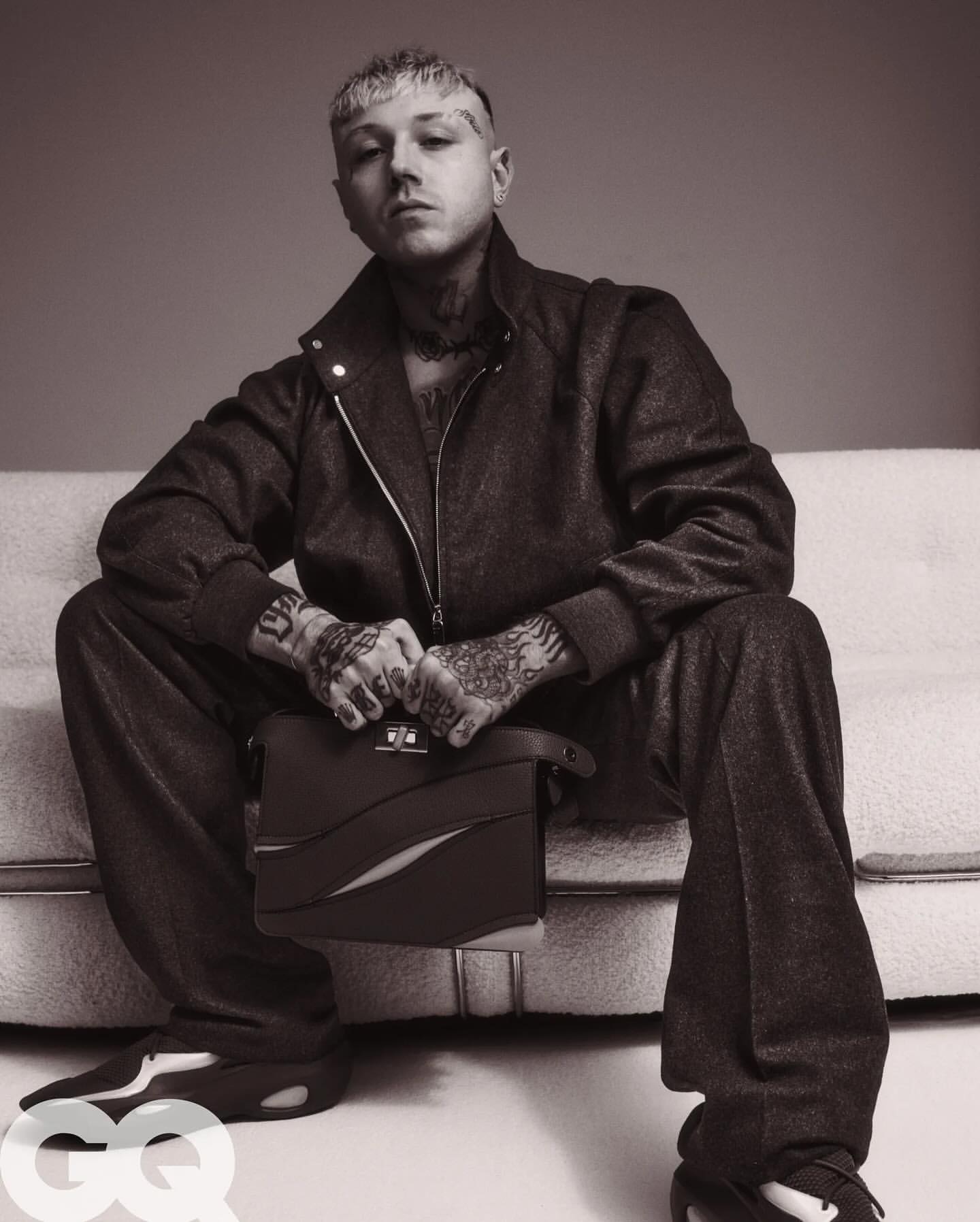 Monochrome portrait of tattooed adult male model in matching jacket and pants, sitting on a sofa holding a leather briefcase.