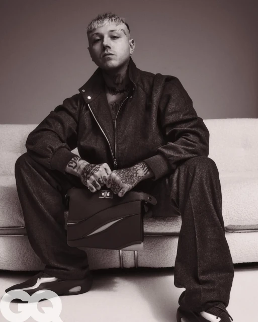 Monochrome portrait of tattooed adult male model in matching jacket and pants, sitting on a sofa holding a leather briefcase.