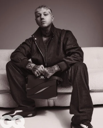 Monochrome portrait of tattooed adult male model in matching jacket and pants, sitting on a sofa holding a leather briefcase.