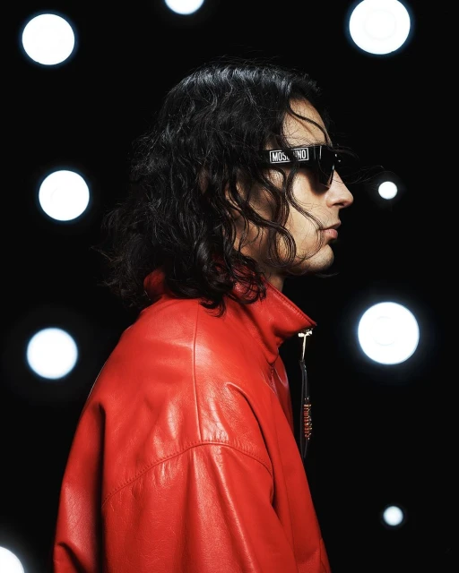 Man in red jacket and sunglasses with a Moschino logo poses against a black background with bright, out-of-focus light orbs.