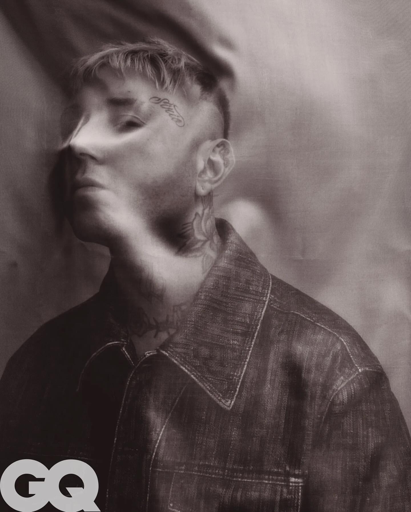 Portrait of a man with tattoos wearing a denim jacket against a draped background in a studio setting, lit with diffused lighting.