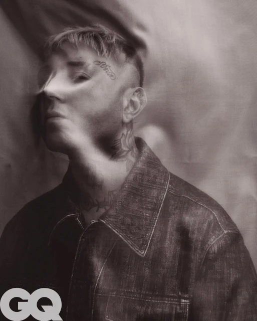 Portrait of a man with tattoos wearing a denim jacket against a draped background in a studio setting, lit with diffused lighting.