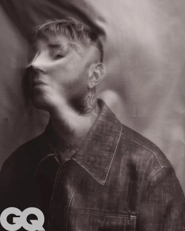 Portrait of a man with tattoos wearing a denim jacket against a draped background in a studio setting, lit with diffused lighting.