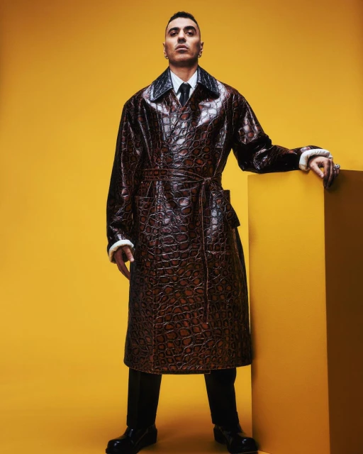 Man posing in a studio wearing a long brown croc-skin pattern coat with fur trim, a white collared shirt, a black tie, and black dress pants.