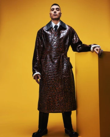 Man posing in a studio wearing a long brown croc-skin pattern coat with fur trim, a white collared shirt, a black tie, and black dress pants.