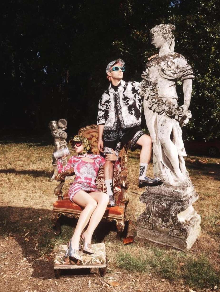 Fashion editorial shot in a garden featuring a man posing next to a statue and a female mannequin seated on a decorative chair.