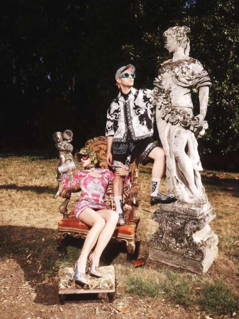 Fashion editorial shot in a garden featuring a man posing next to a statue and a female mannequin seated on a decorative chair.