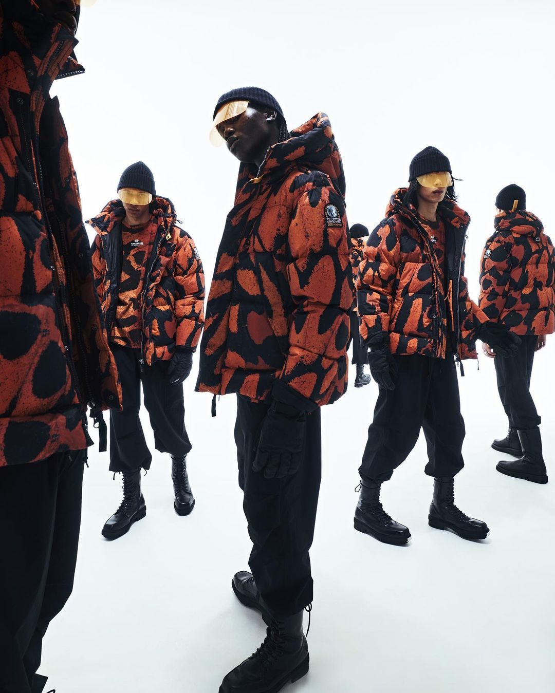 Several people wearing orange and black patterned puffer jackets, black pants, boots and hats stand in a bright studio.
