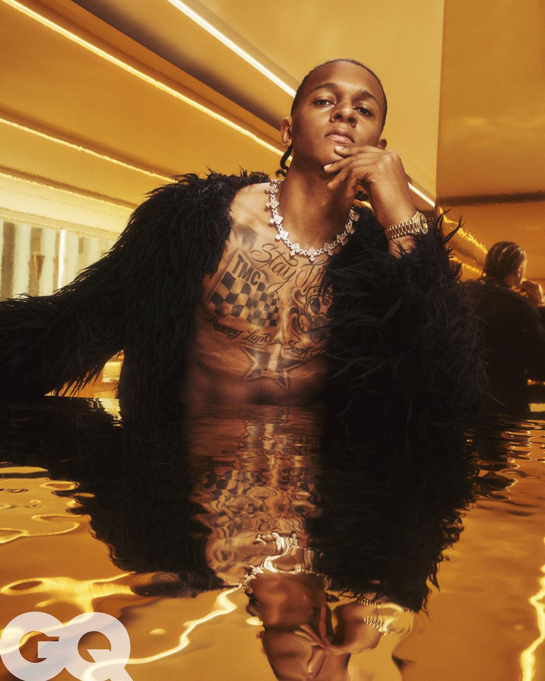 Portrait of a topless Black man with a serious expression in gold-toned water, wearing a coat, jewelry, reflecting with GQ logo.