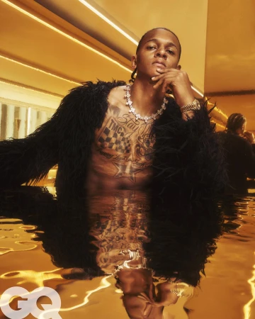 Portrait of a topless Black man with a serious expression in gold-toned water, wearing a coat, jewelry, reflecting with GQ logo.