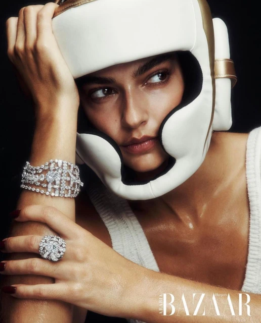 Woman in a boxing head guard poses with diamonds, wrist bracelet, and ring against a black background in a studio shot for Bazaar.