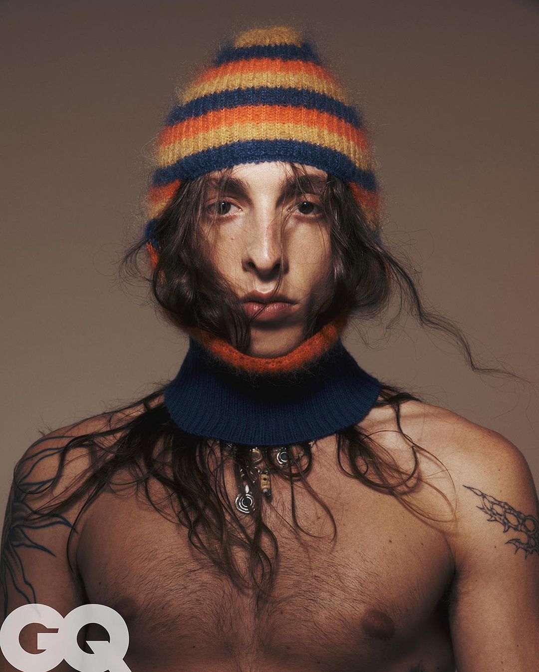 Close-up studio portrait of a shirtless man with long hair wearing a knit beanie hat and collar against a neutral backdrop.