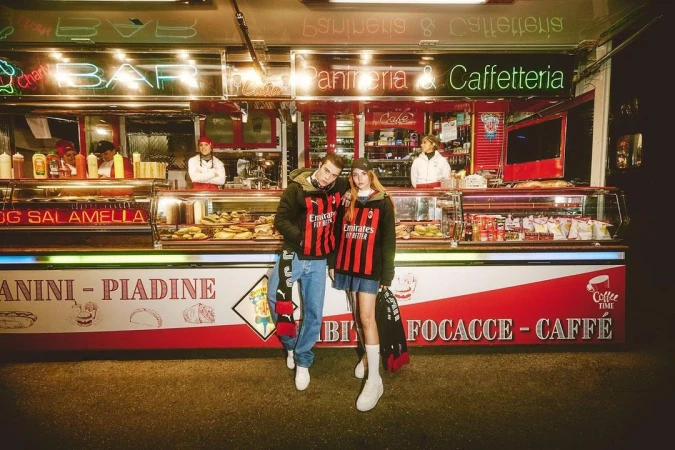 Two people in matching sports jackets stand in front of a street food cafe offering panini and focaccia, with several workers visible inside.
