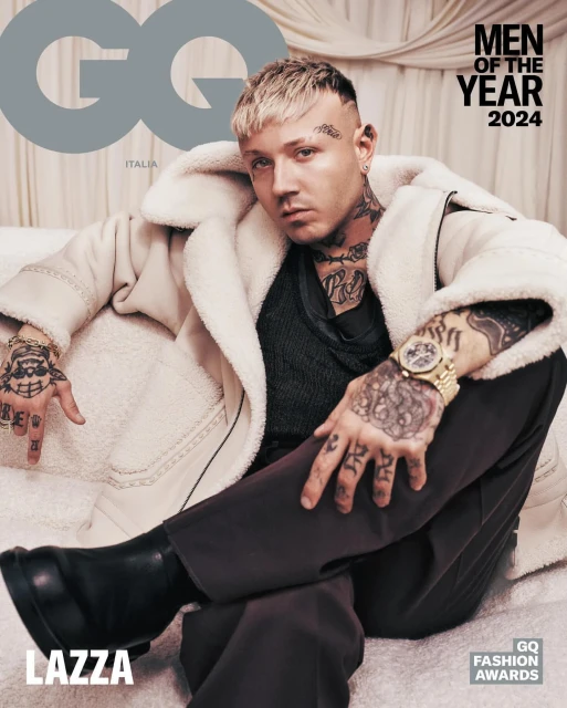 GQ magazine cover featuring platinum-haired model Lazza wearing a cream-colored coat, brown pants, and black boots, sitting on white fabric.