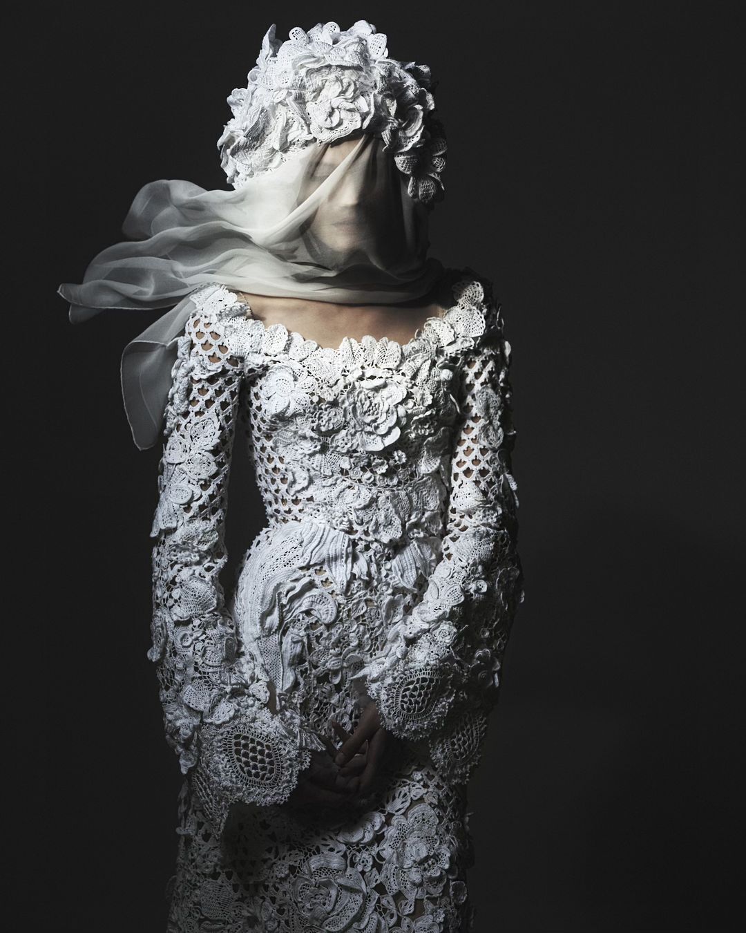 Model wearing an ornate, white, full-length, long-sleeved dress with lace details and a sheer head covering in a studio setting.