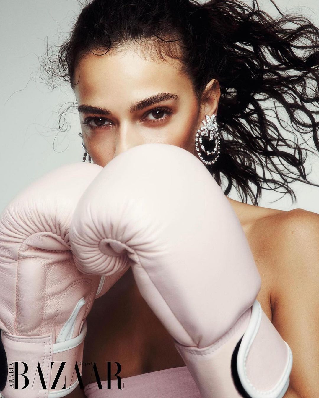Fashion shot of a woman with dark hair wearing statement earrings and pink boxing gloves in a bright, neutral studio environment.