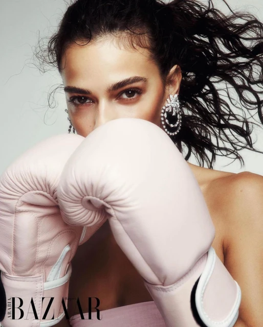 Fashion shot of a woman with dark hair wearing statement earrings and pink boxing gloves in a bright, neutral studio environment.
