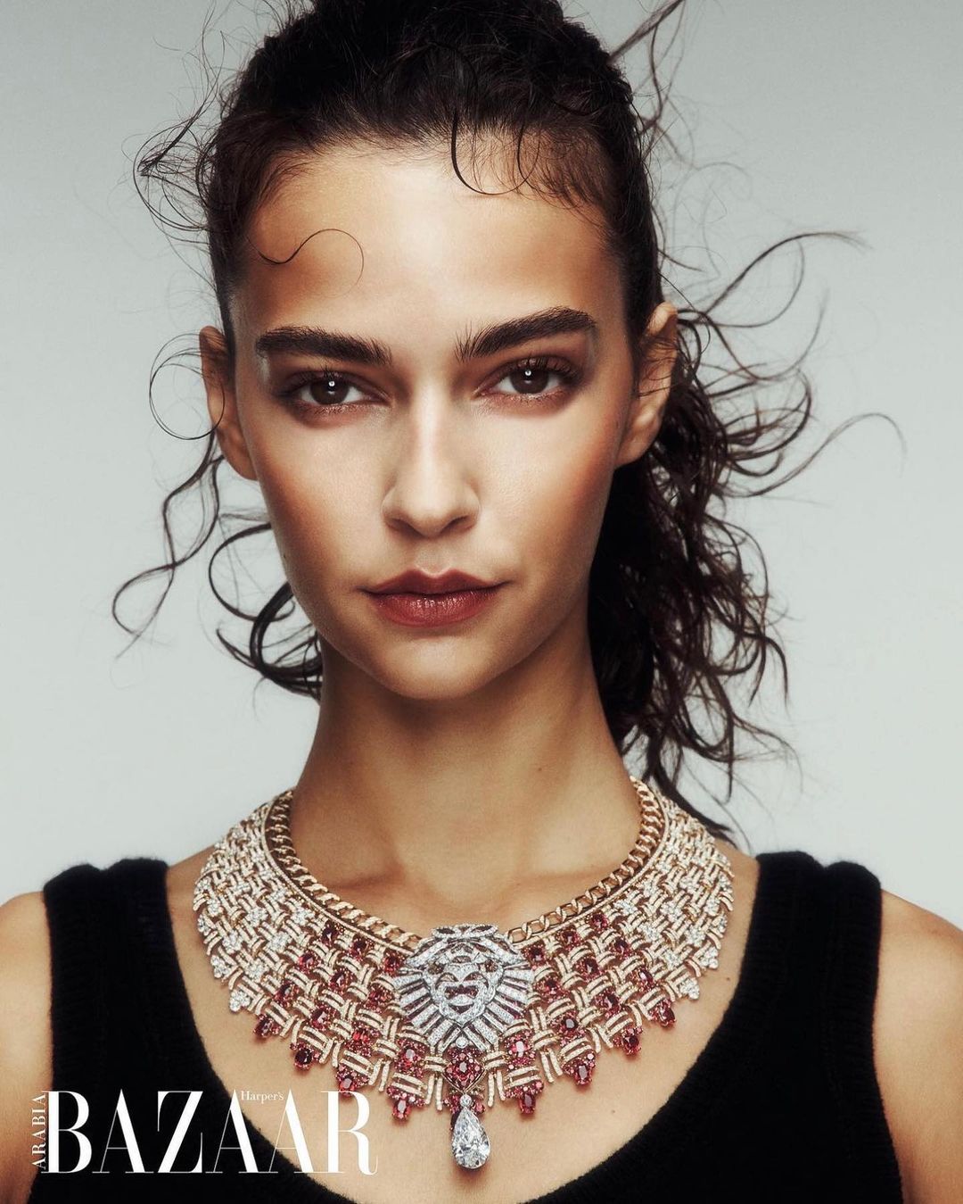 Close-up of a woman with dark hair wearing an ornate diamond and ruby necklace, shot against a light grey background.