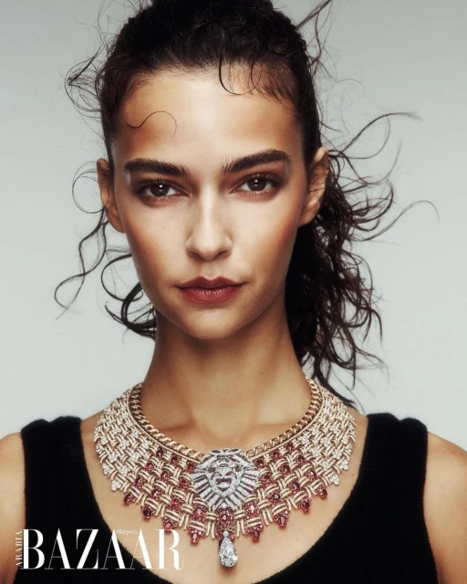 Close-up of a woman with dark hair wearing an ornate diamond and ruby necklace, shot against a light grey background.