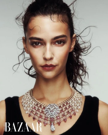 Close-up of a woman with dark hair wearing an ornate diamond and ruby necklace, shot against a light grey background.
