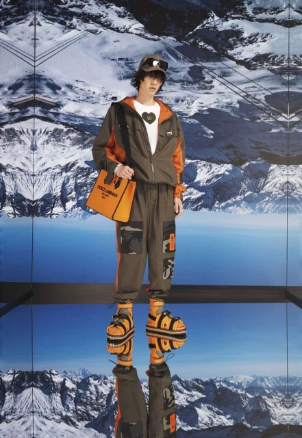 Fashion shot of a male model in olive and orange outdoor wear with a handbag, standing against a mountain backdrop reflection.