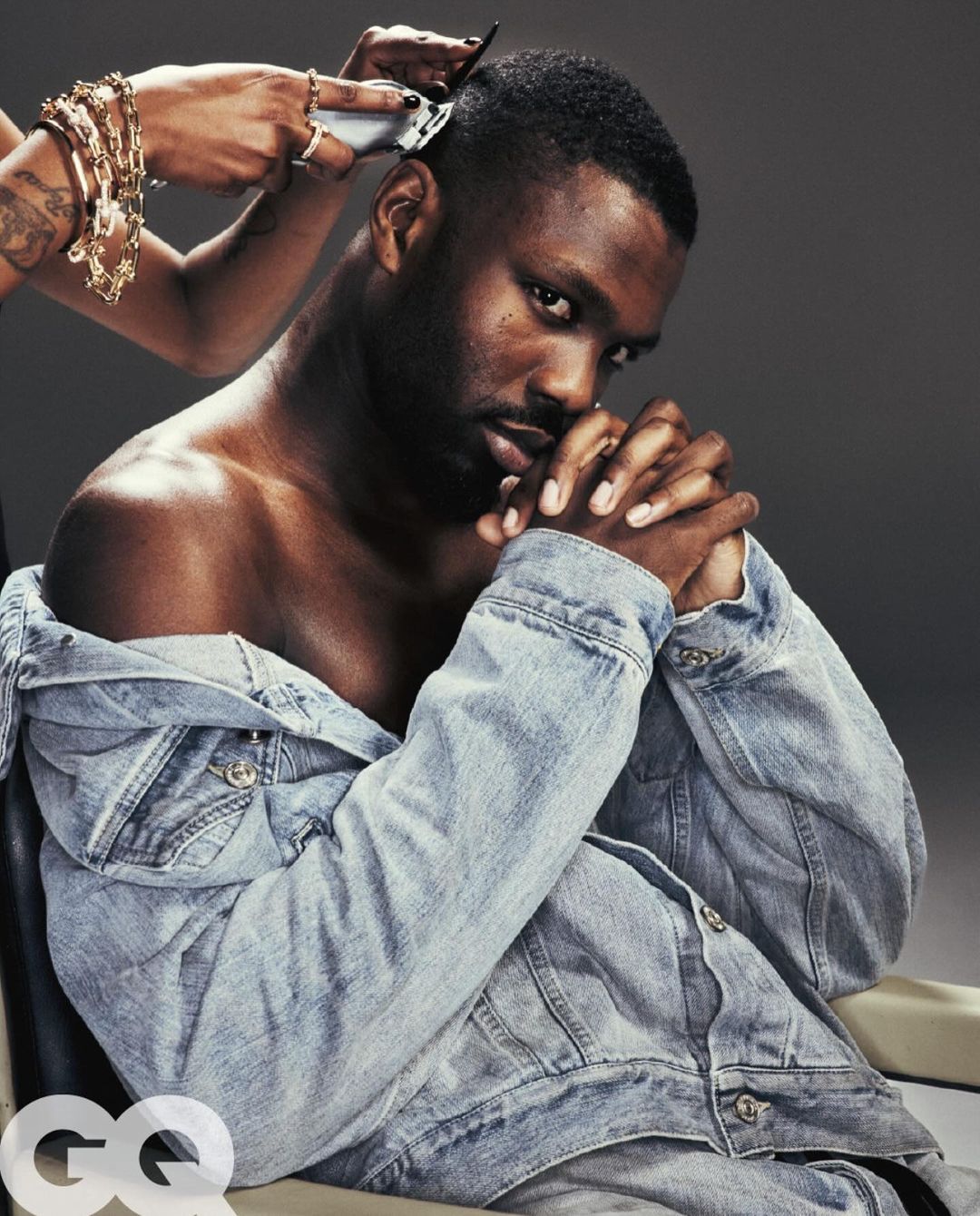 Close-up studio shot of a Black man getting a haircut with an electric razor, wearing a denim jacket, against a grey background.