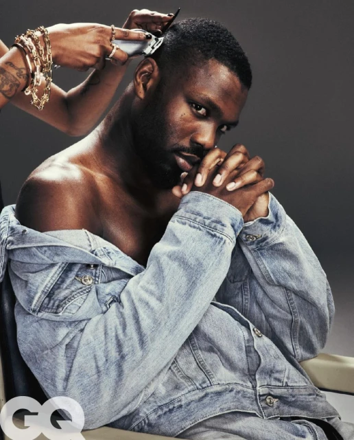 Close-up studio shot of a Black man getting a haircut with an electric razor, wearing a denim jacket, against a grey background.